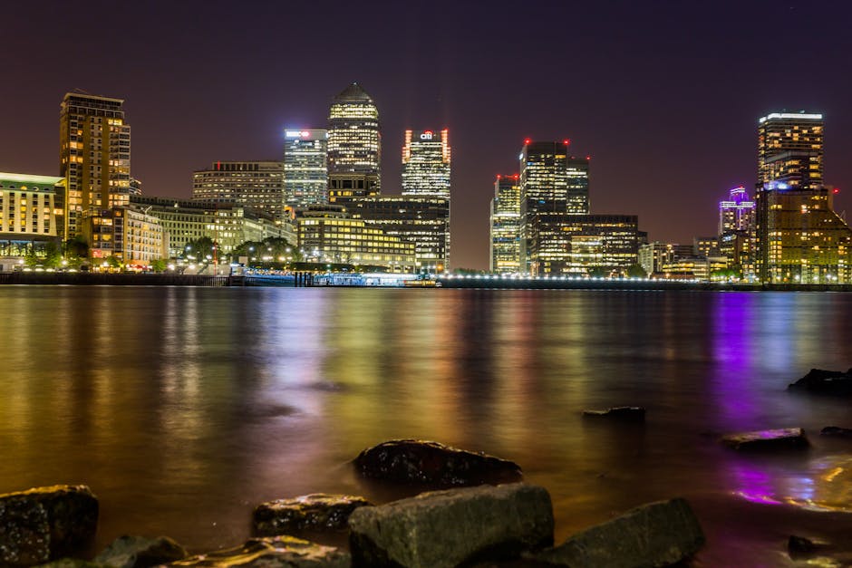 Nighttime view of Canary Wharf's skyline from across the River Thames, featuring illuminated high-rise office buildings made of glass and steel, reflecting colorful city lights onto the water. The sce