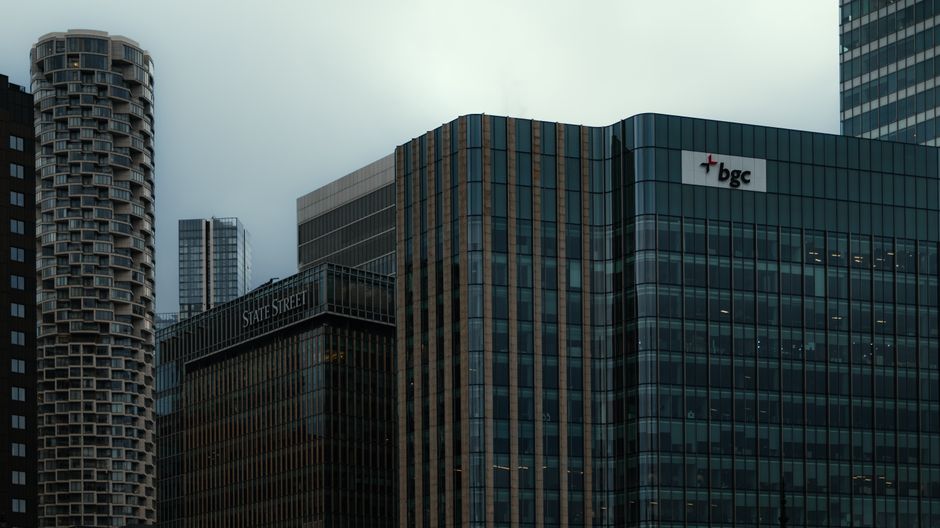 Nighttime view of Canary Wharf's skyline from across the River Thames, featuring illuminated high-rise office buildings made of glass and steel, reflecting colorful city lights onto the water. The sce