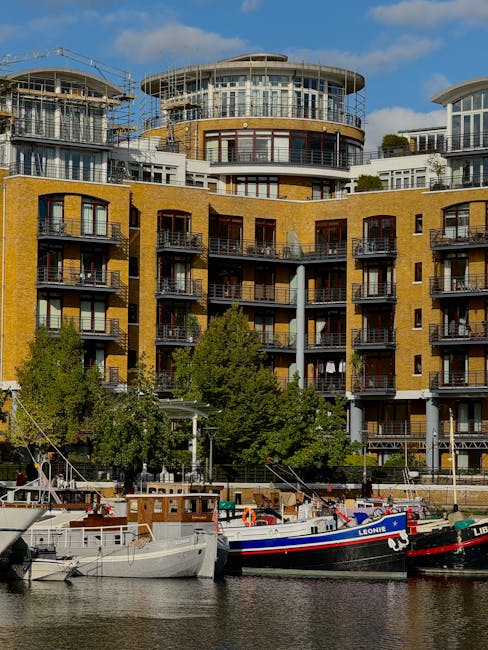 A canal lined with modern residential buildings made of beige brick and dark metal accents, with large windows and small balconies, reflecting in the calm water below. The scene is illuminated by natu